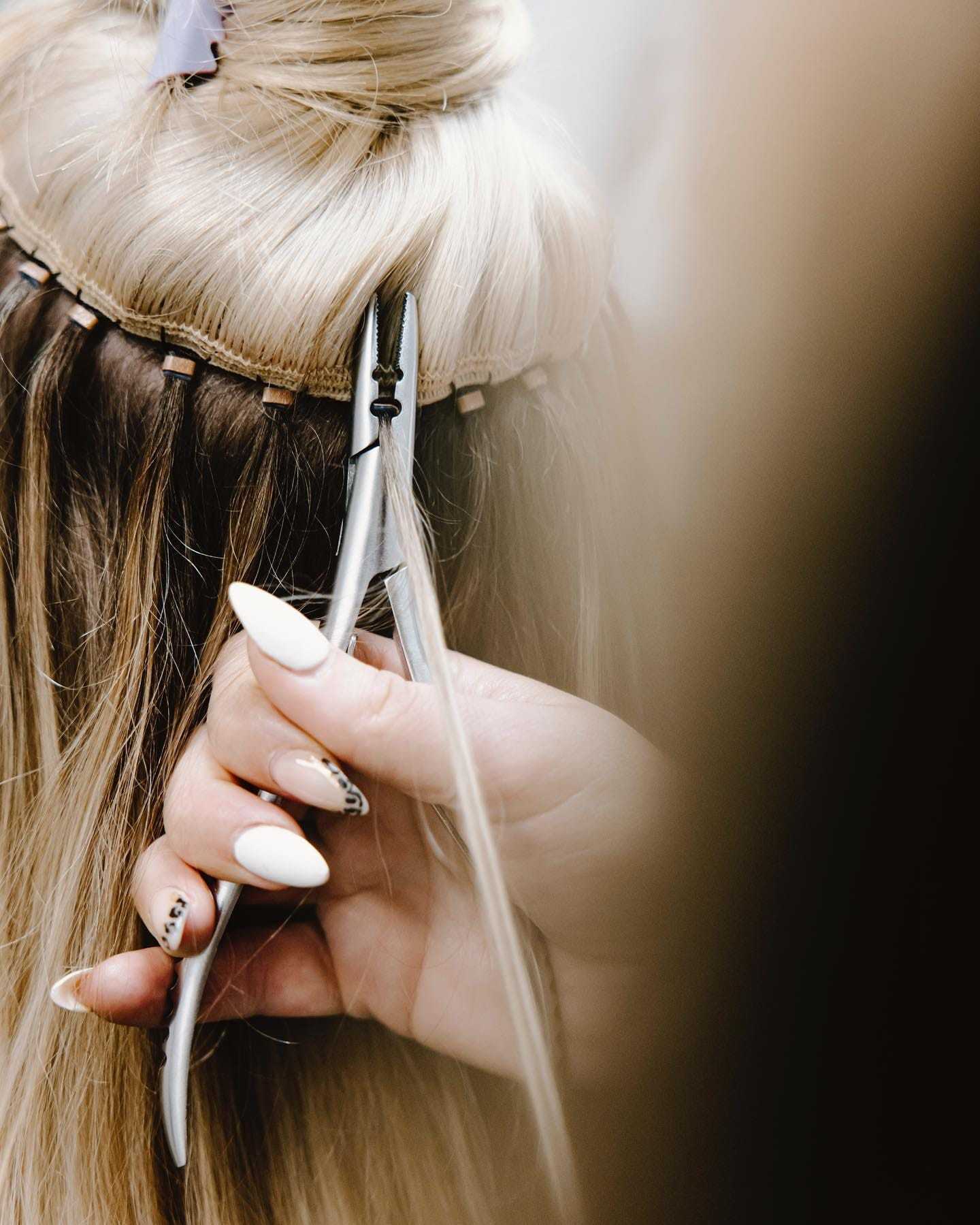 Hair extensions being applied to long blonde hair with a tool in a salon setting.