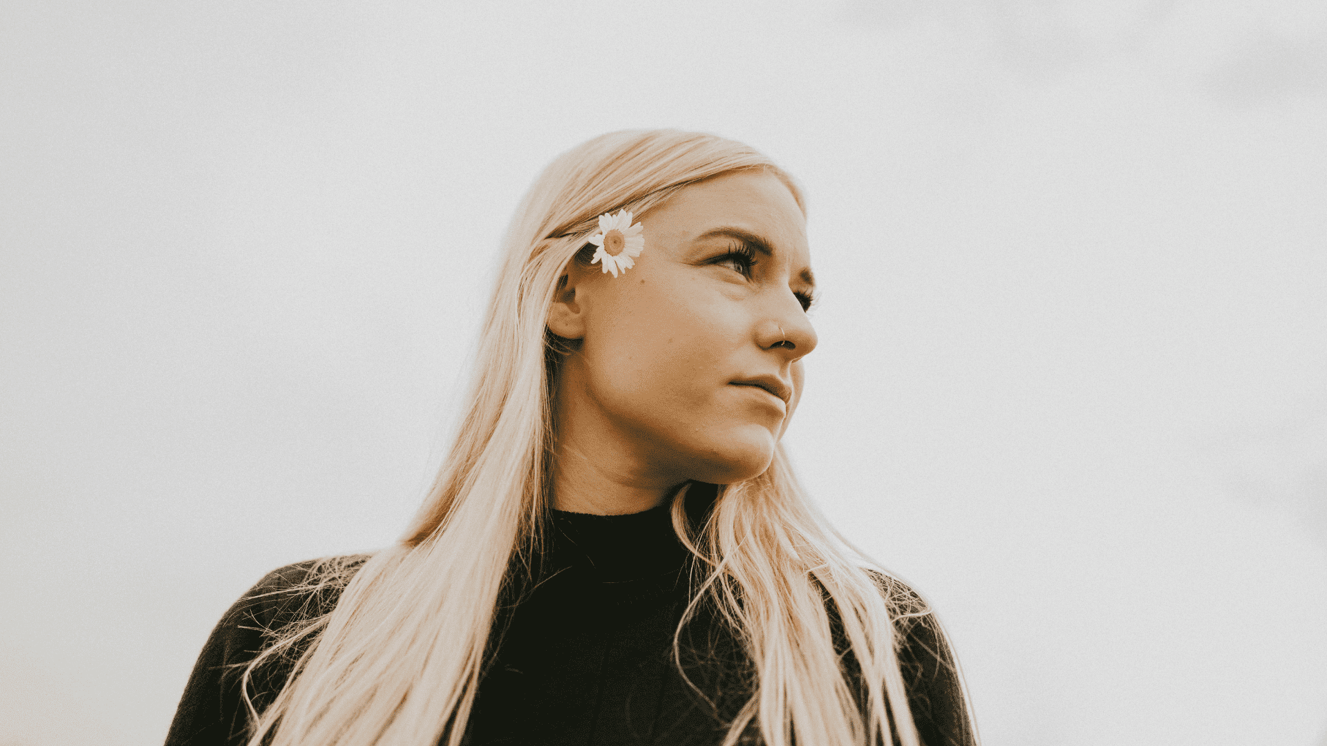 Blonde woman with a daisy in hair, gazing to the side, set against a light sky background.